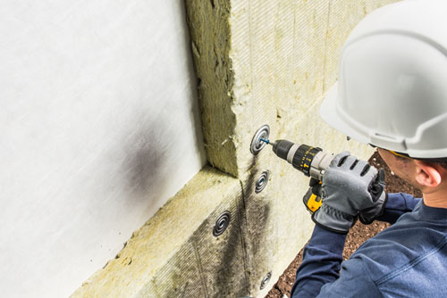 Woodman Insulation installer using a power drill to secure mineral wool insulation panels to an interior wall during a commercial project.