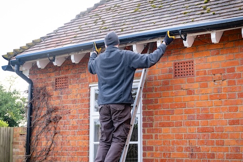 Man on a ladder performing gutter replacement on a brick house with asphalt roof