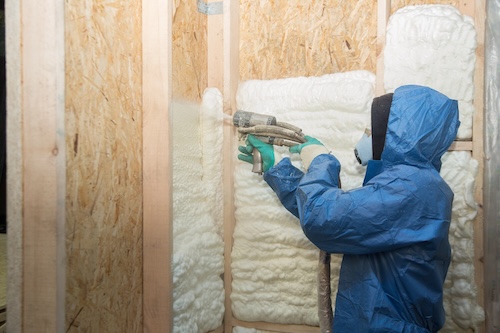 worker in blue protective suit sprays foam insulation into new wall cavity