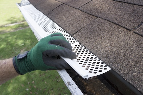 closeup of hand wearing green work gloves placing mesh gutter guard onto end of asphalt roof