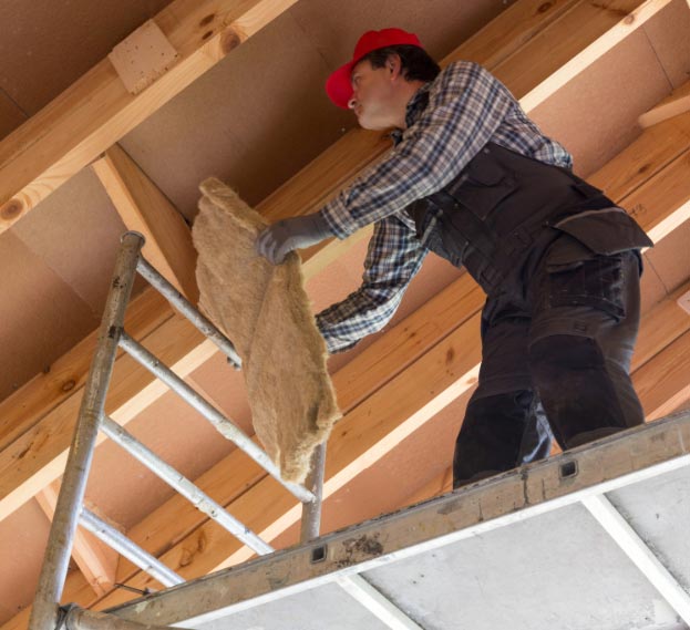 A worker on scaffolding installing mineral wool insulation under the roof of a house, wearing protective gloves and a red cap.