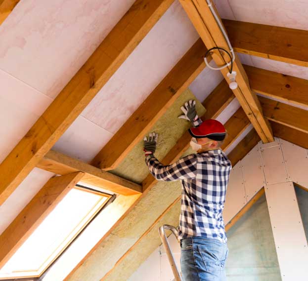 A worker installing mineral wool insulation in an attic space, wearing a plaid shirt, gloves, and a protective mask.