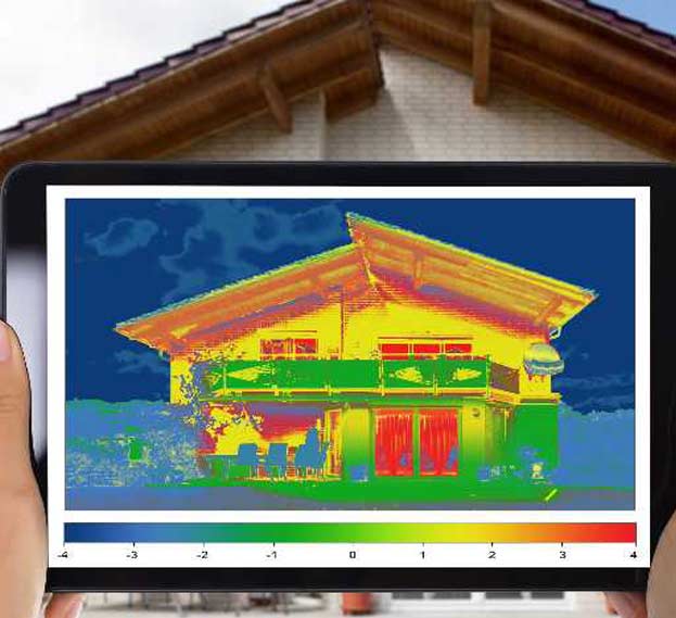 A person holds a tablet in front of a house, displaying a thermal imaging scan of the building. The thermal image shows various heat levels, with red and yellow indicating warmer areas and green and blue representing cooler areas. The house in the background has a wooden roof and white brick walls.