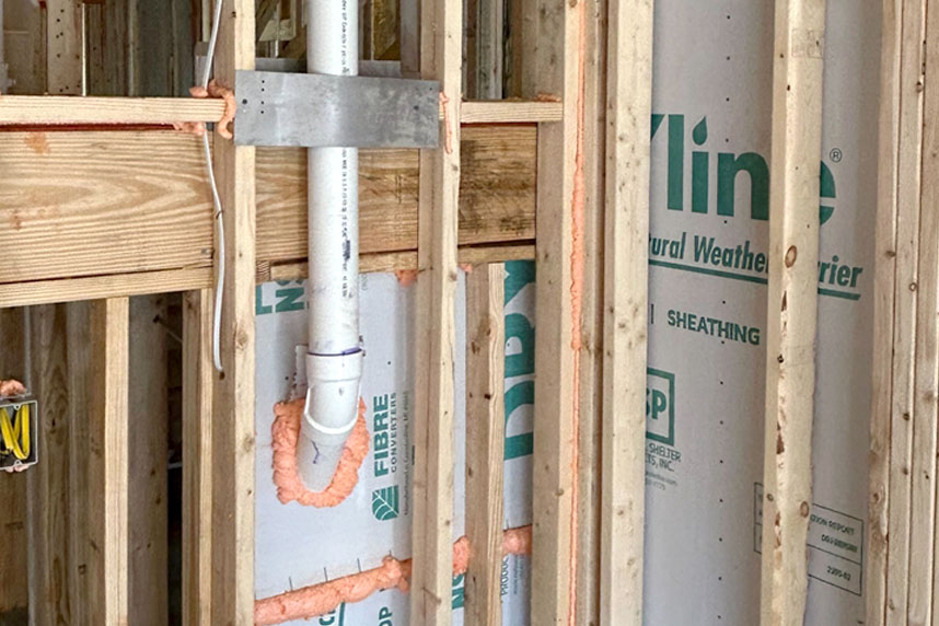 Close-up of spray foam sealant applied around a PVC pipe and sheathing during new home construction.
