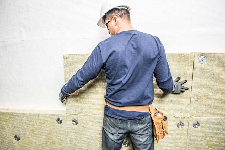 Installer placing mineral wool boards against a vertical exterior wall with fasteners, viewed from the back.