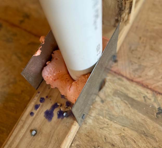Close-up of a white pipe sealed with spray foam and protected by a metal nail guard where it passes through floor framing.