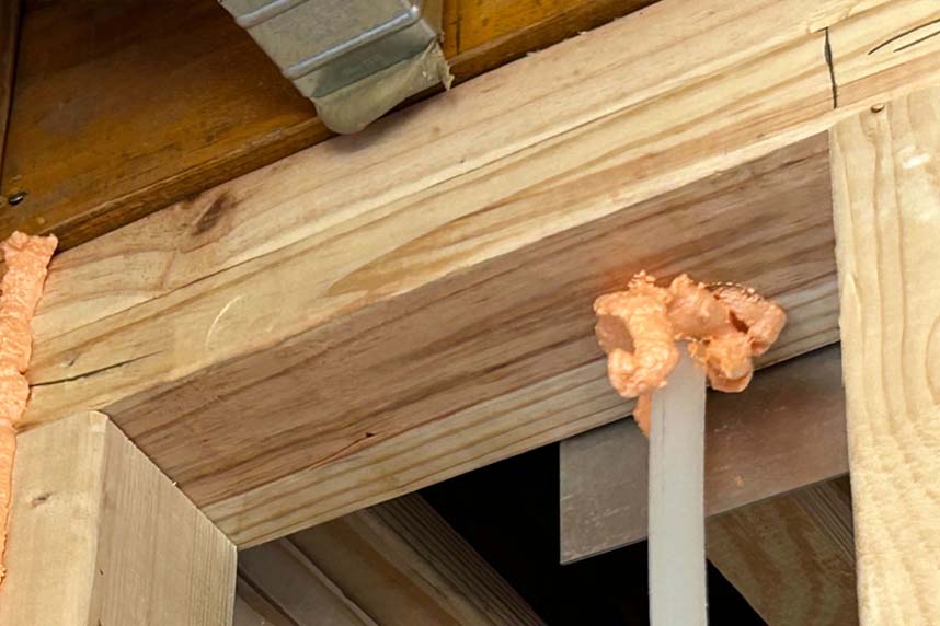 Spray foam applied around a single wire penetration through wood framing at the ceiling level of a residential structure.