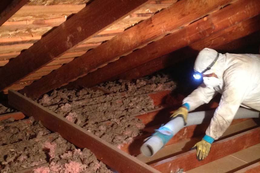 Worker in protective suit and headlamp removing old insulation from an attic using a vacuum hose.