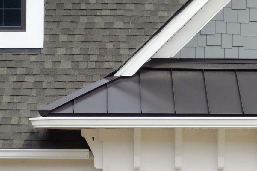 Close-up of a residential roof showing asphalt shingles, gray metal roofing detail, and white trim around a window.